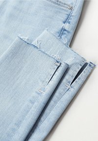 Jeans de mezclilla azul claro con un dobladillo deshilachado y un detalle de abertura cerca del tobillo, sobre un fondo blanco.