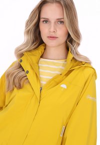 Żółta wodoodporna kurtka z kapturem, z małym białym logo parasola i guzikami, noszona na żółto-białym paskowanym topie.
