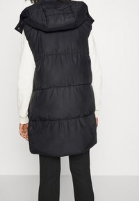 Svart pufferväst med huva, med kviltade sektioner och en lös passform, bärs över en vit långärmad tröja och svarta texturerade leggings.