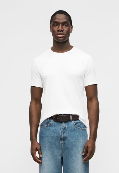 Homme debout face à l'avant, portant un t-shirt blanc à manches courtes uni et un jean bleu avec une ceinture marron foncé, sur un fond uni.