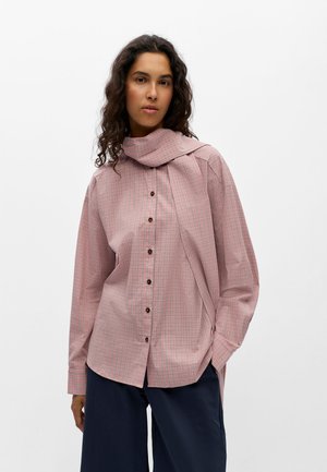 Femme aux cheveux bouclés foncés porte une chemise à carreaux rose avec boutons, col écharpe prolongé, et pantalon large bleu marine.