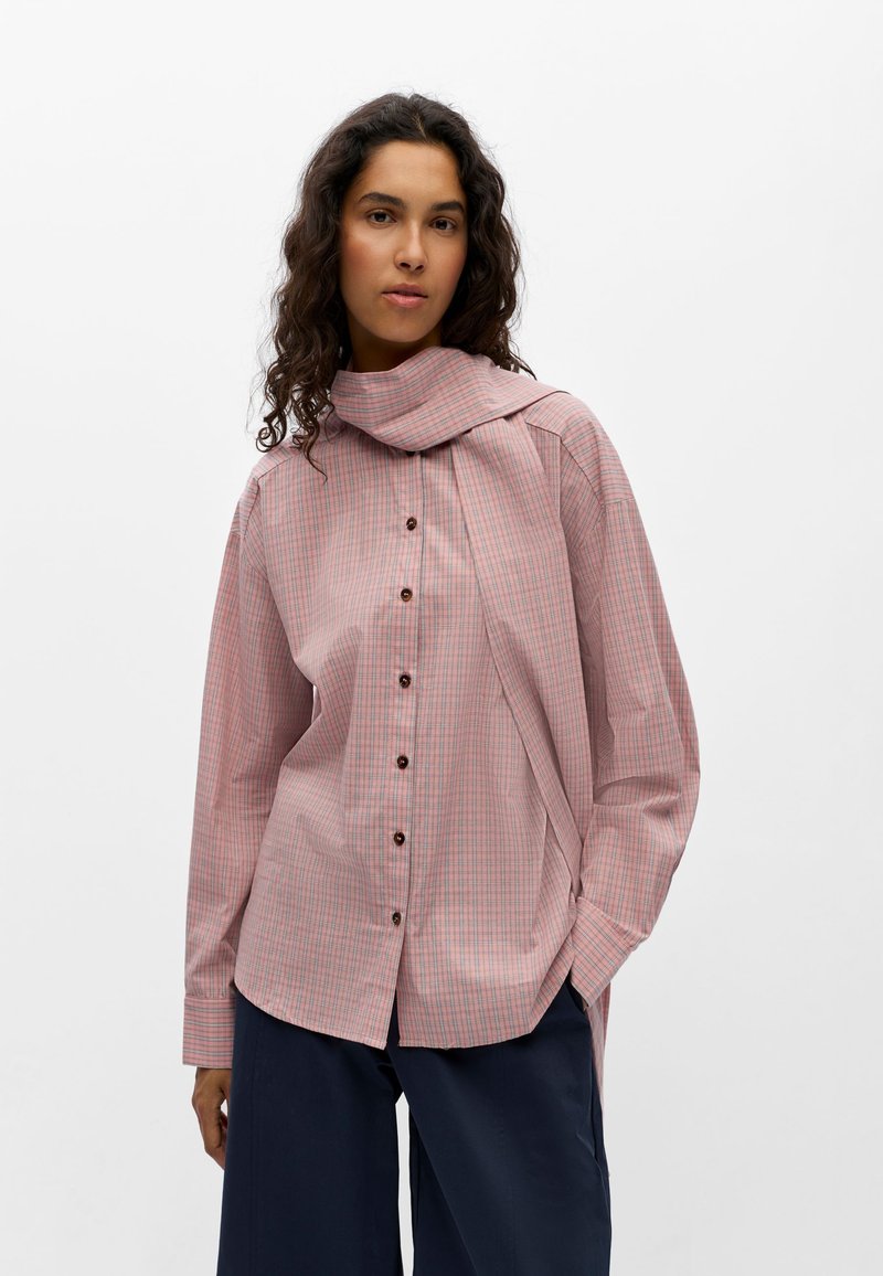 Femme aux cheveux bouclés foncés porte une chemise à carreaux rose avec boutons, col écharpe prolongé, et pantalon large bleu marine.