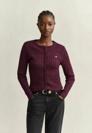 Junge Frau mit geflochtenem Haar, die eine weinrote Zopfmuster-Strickjacke und schwarze Jeans mit schwarzem Gürtel trägt, steht mit den Händen in den Taschen.