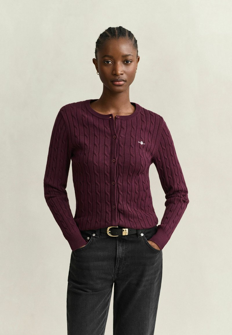 Jeune femme aux cheveux tressés portant un cardigan bordeaux en tricot torsadé et un jean noir avec une ceinture noire, debout les mains dans les poches.