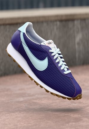 Lilla og lyseblå Nike løbesko med hvide snørebånd og gummisål, vist svævende over en glat udendørs overflade.