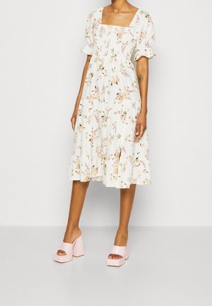 Femme portant une robe midi blanche à fleurs avec manches bouffantes et encolure carrée, assortie à des mules roses à semelle compensée.
