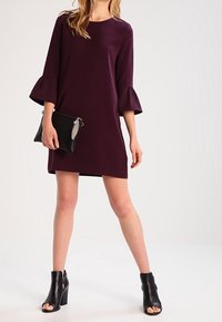 Kvinna i en plommonfärgad knälång klänning med trumpetärmar, hållande en svart clutch och iförd svarta öppna ankelstövlar.