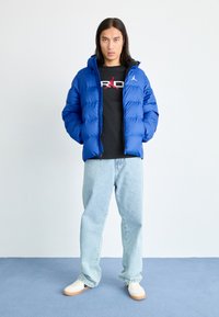 Blå puffjakke med hette, med en blank overflate og quiltet design. Bæres over en svart T-skjorte og lyseblå jeans, sammen med hvite sko.