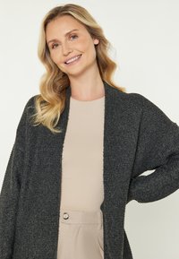 Cardigan texturat negru cu deschidere în față și mâneci largi, purtat peste un top bej deschis. Prezentă o croială lejeră și un material moale.