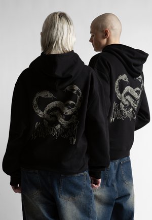 Dvaja ľudia v čiernych mikinách s kapucňou s prepletenými hadími motívmi a štýlizovaným textom na chrbte, v kombinácii s vyblednutými modrými džínsami.