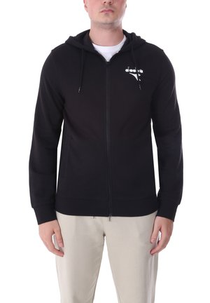 Uomo che indossa una felpa nera con zip e logo bianco sul petto e pantaloni beige, in piedi davanti a uno sfondo bianco.