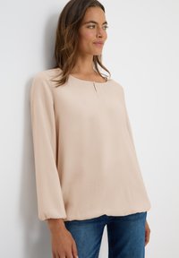 Vrouw in een lichtbeige blouse met lange mouwen en een klein knoopdetail bij de halslijn, gecombineerd met een blauwe jeans, staand tegen een witte muur.