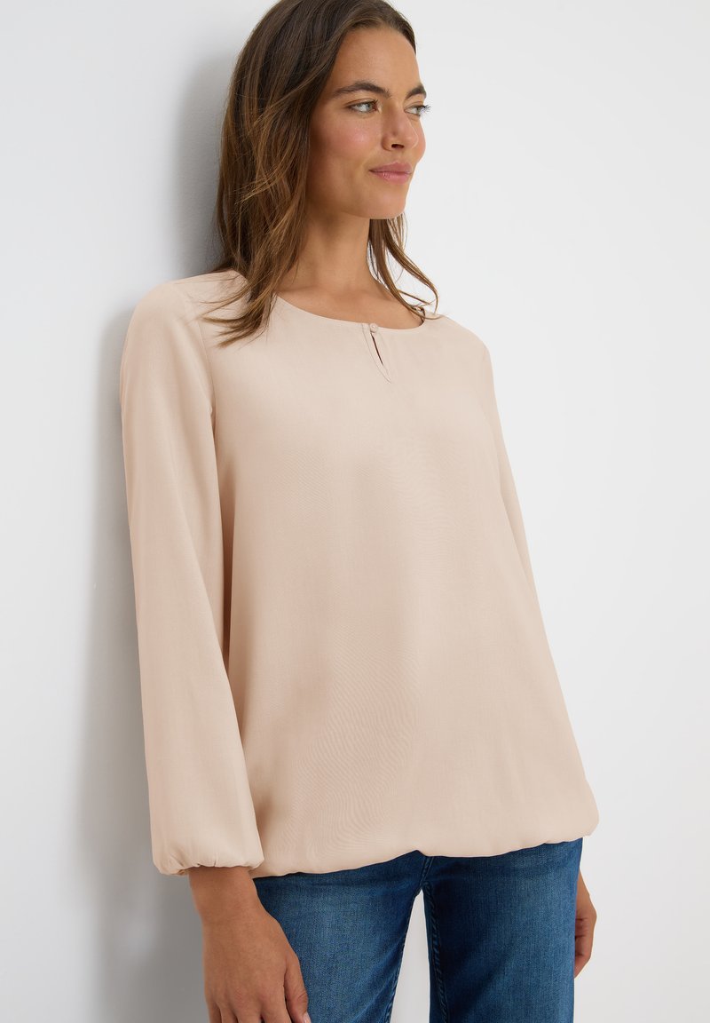 Vrouw in een lichtbeige blouse met lange mouwen en een klein knoopdetail bij de halslijn, gecombineerd met een blauwe jeans, staand tegen een witte muur.