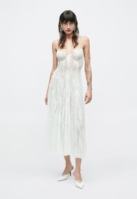 Áttetsző fehér ruha virágos hímzéssel, testhez simuló felsőrésszel és spagetti pántokkal. A lágyan hullámzó szoknya középhosszú, fehér magassarkú cipővel párosítva.