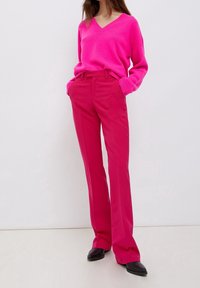 Fuchsiový pletený svetr s výstřihem do V a dlouhými rukávy, doplněný o fuchsiové kalhoty rovného střihu. Oba kusy mají hladkou texturu. Černé boty.