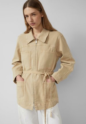 Femme portant une veste beige zippée avec de grandes poches frontales et une ceinture nouée à la taille, posant les mains dans les poches sur un fond gris.