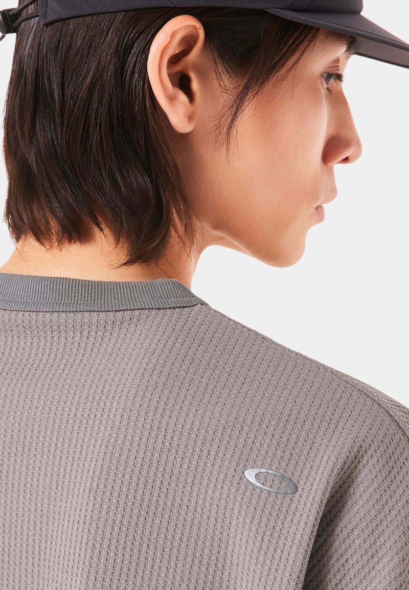 Gray textured shirt with a ribbed design and a round neckline, featuring a small silver logo on the back. Cap worn on the head.