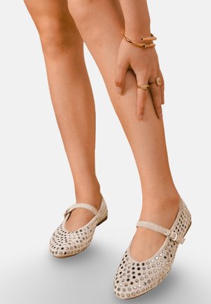 Jambes portant des chaussures Mary Jane beiges avec des clous métalliques circulaires, main reposant sur la jambe, ornée de bagues et de bracelet en or.