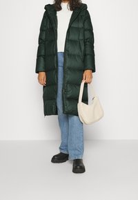 Person med lang, mørkegrøn pufferjakke, hvid sweater, blå jeans, sorte støvler og som holder en lille hvid, blød håndtaske.