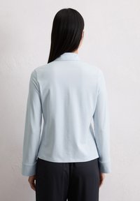Himmelblaues Langarmshirt mit lockerer Passform, Kragen und Nahtdetails auf der Rückseite. Glatte Textur mit einem dezenten Glanz. Kombiniert mit dunklen Hosen.
