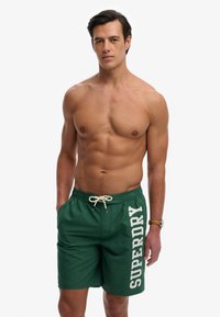 Homme musclé aux cheveux foncés portant un short de bain vert avec le texte blanc "SUPERDRY" et une montre, debout devant un fond clair uni.