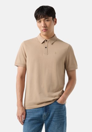Junger Mann trägt ein beiges kurzärmeliges Poloshirt und blaue Jeans mit einer Hand in der Tasche vor einfachem Hintergrund.