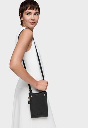 Kobieta w białej sukience bez rękawów, niosąca małą czarną torebkę na pasku na ramię z złotym ozdobnym zawieszką i tłoczonym nazwiskiem marki.