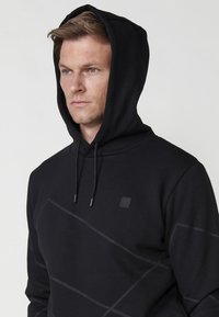 Hanorac negru cu glugă ajustabilă, având un model geometric subtil și o etichetă cu brand pe piept. Textură netedă, croială casual.