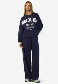 Jūras zilas krāsas sporta krekls un saskaņotas bikses, kas izgatavotas no mīksta auduma, ar "BOROUGH Market" grafiku. Brīvs piegriezums ar nolaižamām plecu šuvēm un regulējamu jostu.