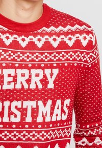 Rode gebreide kersttrui met witte tekst "MERRY CHRISTMAS", geometrische patronen en een geribde halslijn.