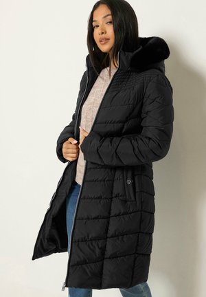Femme portant un long manteau noir matelassé avec une capuche bordée de fourrure, un pull beige et un jean bleu sur un fond clair uni.
