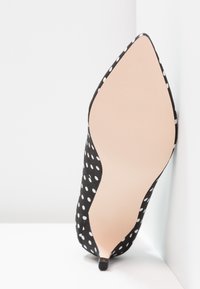 Pointed-toe black high heel with white polka dots, viewed from sole, positioned against white wall and floor corner.