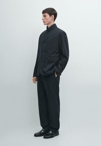 Sort jakke med knapper foran, stående krave, sidelommer og løs pasform, kombineret med mørke vidde bukser og sorte loafers.