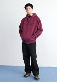Felpa con cappuccio bordeaux con logo, pantaloni neri e sneakers a fantasia. Il tessuto sembra morbido; la vestibilità è rilassata. Sfondo bianco.