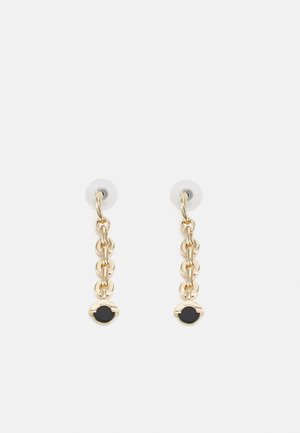 Boucles d'oreilles pendantes en chaîne dorée avec accents circulaires noirs, présentées sur un fond gris clair uni.