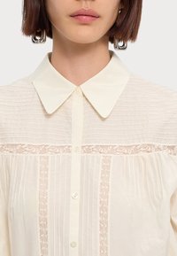 Cream-colored blouse featuring vertical pinstripes, a lace trim, and a classic collar with buttons down the front. Smooth texture.