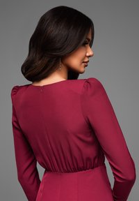 Robe bordeaux avec manches bouffantes, corsage ajusté et taille froncée. Dispose d'une fermeture éclair au dos et d'une texture de tissu lisse et douce.