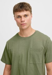 Camiseta de manga corta verde olivo en tejido ligero con cuello redondo y un bolsillo en el pecho. Textura suave y corte relajado.
