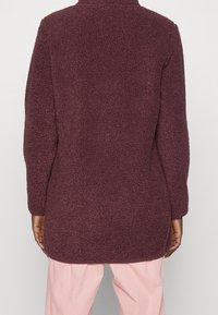 Bordová fleecová bunda s texturovaným, měkkým povrchem; dlouhé rukávy a prodloužený zadní lem, bez viditelných zapínání nebo vzorů.