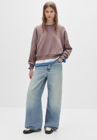 Felpa viola con spalle scese indossata sopra una camicia azzurra a strati, abbinata a jeans larghi blu sbiaditi e scarpe bianco e nere.