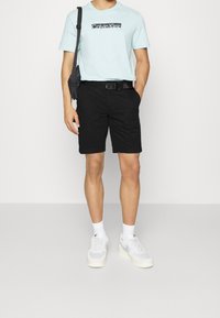 Světle modré tričko s černým logem, černé šortky, bílé kotníkové ponožky a šedé tenisky. Černá ramenní taška a opasek dotvářejí celý outfit.