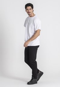 Mann in einem weißen Grafik-T-Shirt, schwarzer Hose und schwarzen Sneakers, der steht und nach vorne schaut, vor einem schlichten hellen Hintergrund.