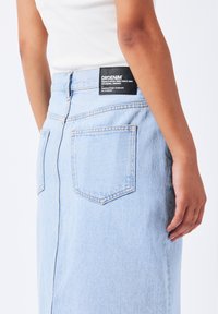 Falda de mezclilla azul clara con dos bolsillos traseros, cintura alta y costuras en relieve. Presenta una etiqueta negra rectangular en la parte trasera con los detalles de la marca.