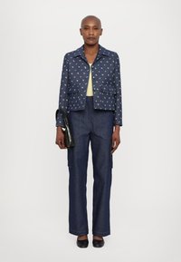 Navy blauwe jacket met bloemengebrocheerde details, gecombineerd met donkere denim wijde broek. Houdt een kleine groene tas met krokodillenpatroon vast.