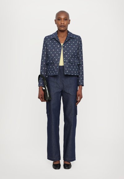 Jachetă bleumarin cu broderie florală, asociată cu pantaloni largi din denim închis. Ține o geantă mică cu model de crocodil verde.