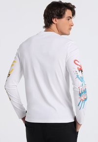 White long-sleeve t-shirt featuring colorful graphic prints in yellow, red, and blue on the sleeves, made of a soft cotton material.