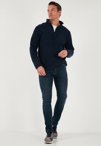 Pullover in fleece blu navy con zip a metà, dal texture morbido, abbinato a jeans denim scuri e sneakers in camoscio grigio con dettagli blu navy.