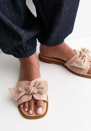 Pieds portant des sandales beige en daim à enfiler avec de grands nœuds noués et des bouts carrés, associées à un pantalon jogger en denim foncé.
