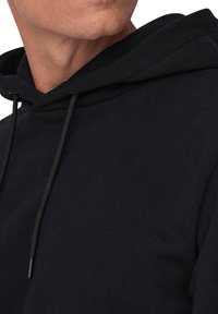 Hoodie in cotone nero con coulisse al collo, texture morbida e vestibilità rilassata. Il tessuto appare leggermente peloso.