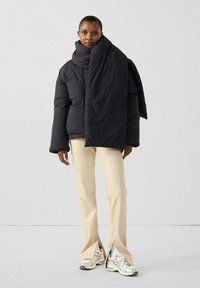 Zwarte pufferjas met oversized ontwerp en unieke sjaalfunctie, gecombineerd met crèmekleurige broek en zilveren sneakers. Gladde texturen duidelijk zichtbaar.